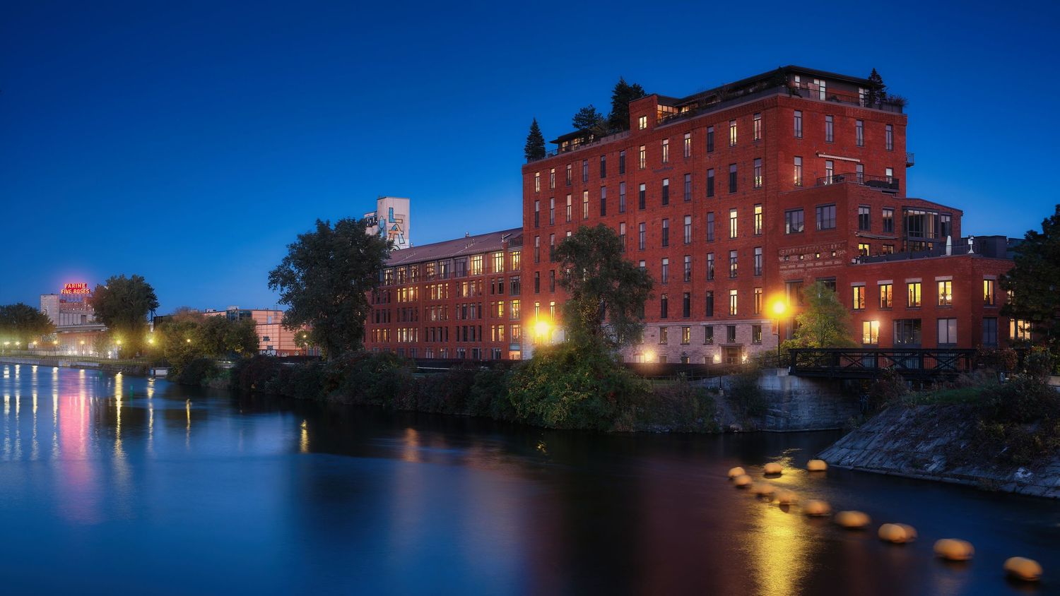 Historic red brick mill building reflected in a calm river at blue hour twilight with city lights glowing in background.