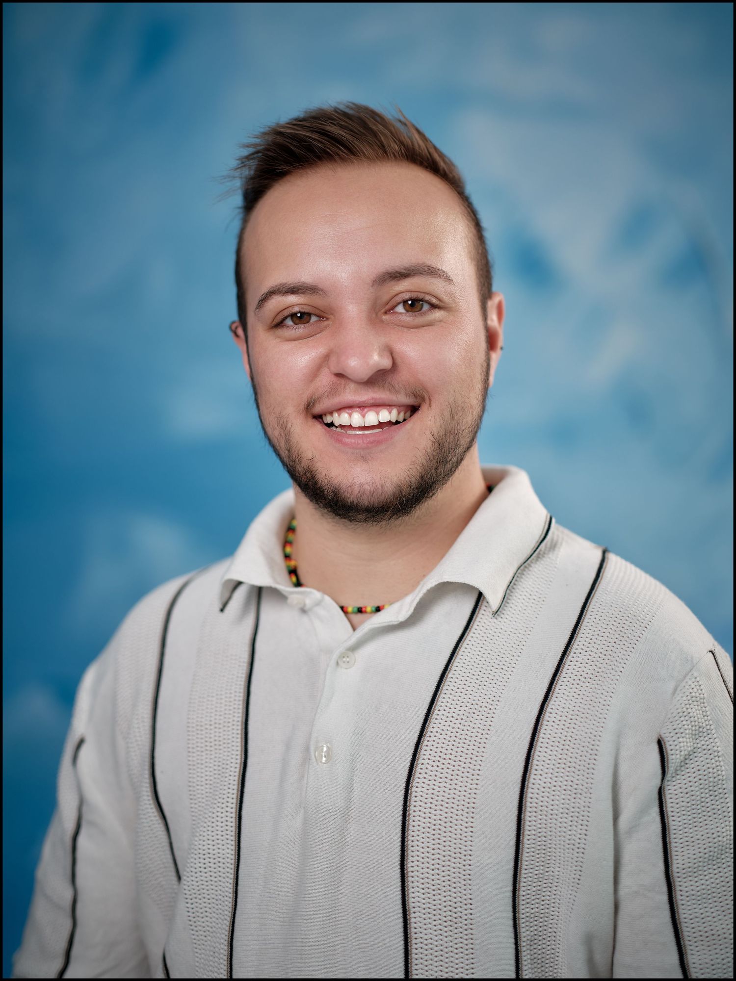 Smiling young man with short hair and beard wearing a striped polo shirt against a blue background.