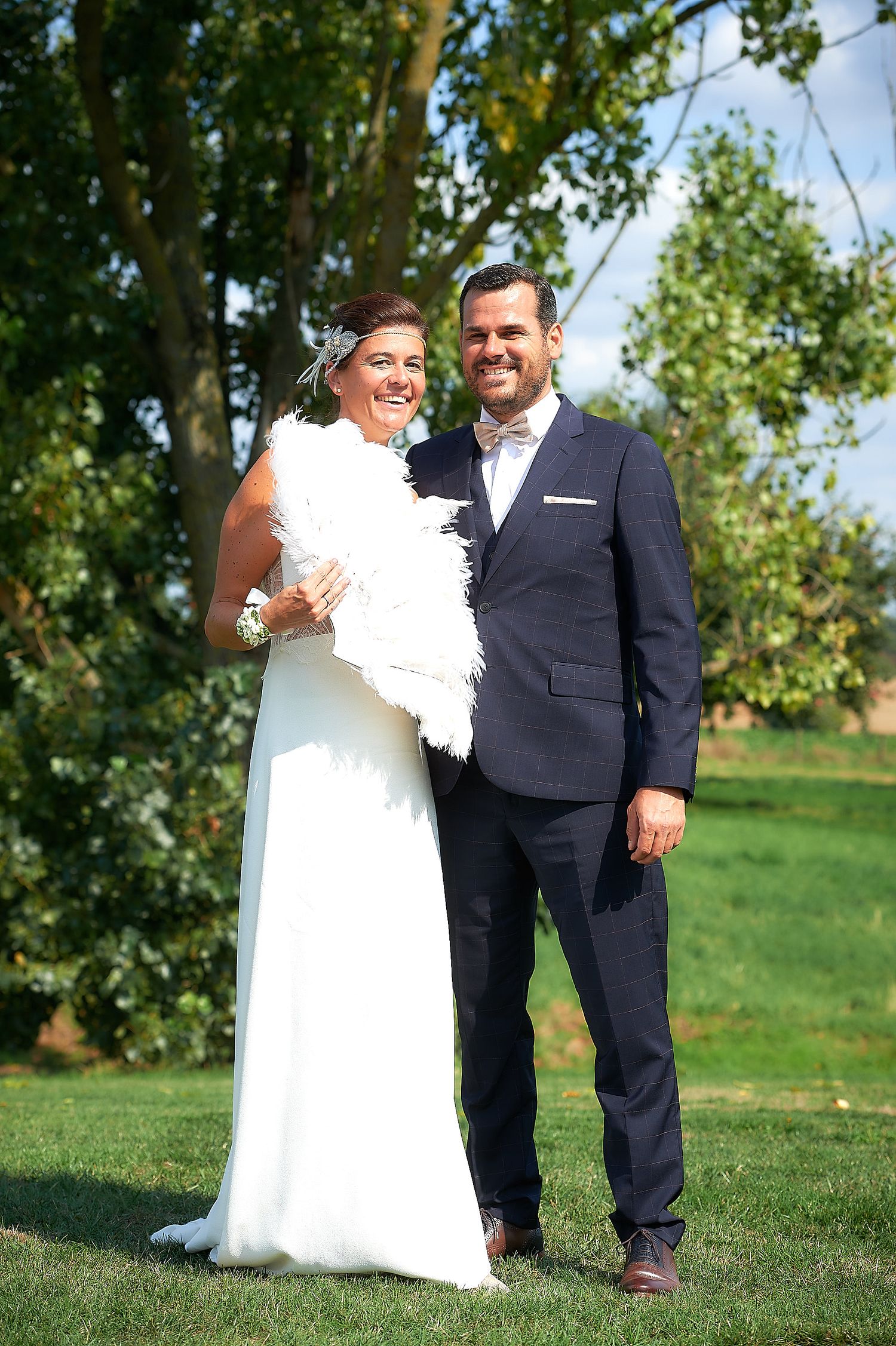 Happy bride and groom posing outdoors on green grass, bride in white gown with feather wrap, groom in navy suit.