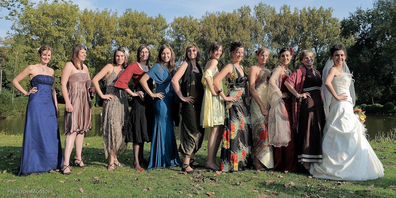 A group of women in elegant dresses pose outdoors near a pond, including a bride in a white gown.