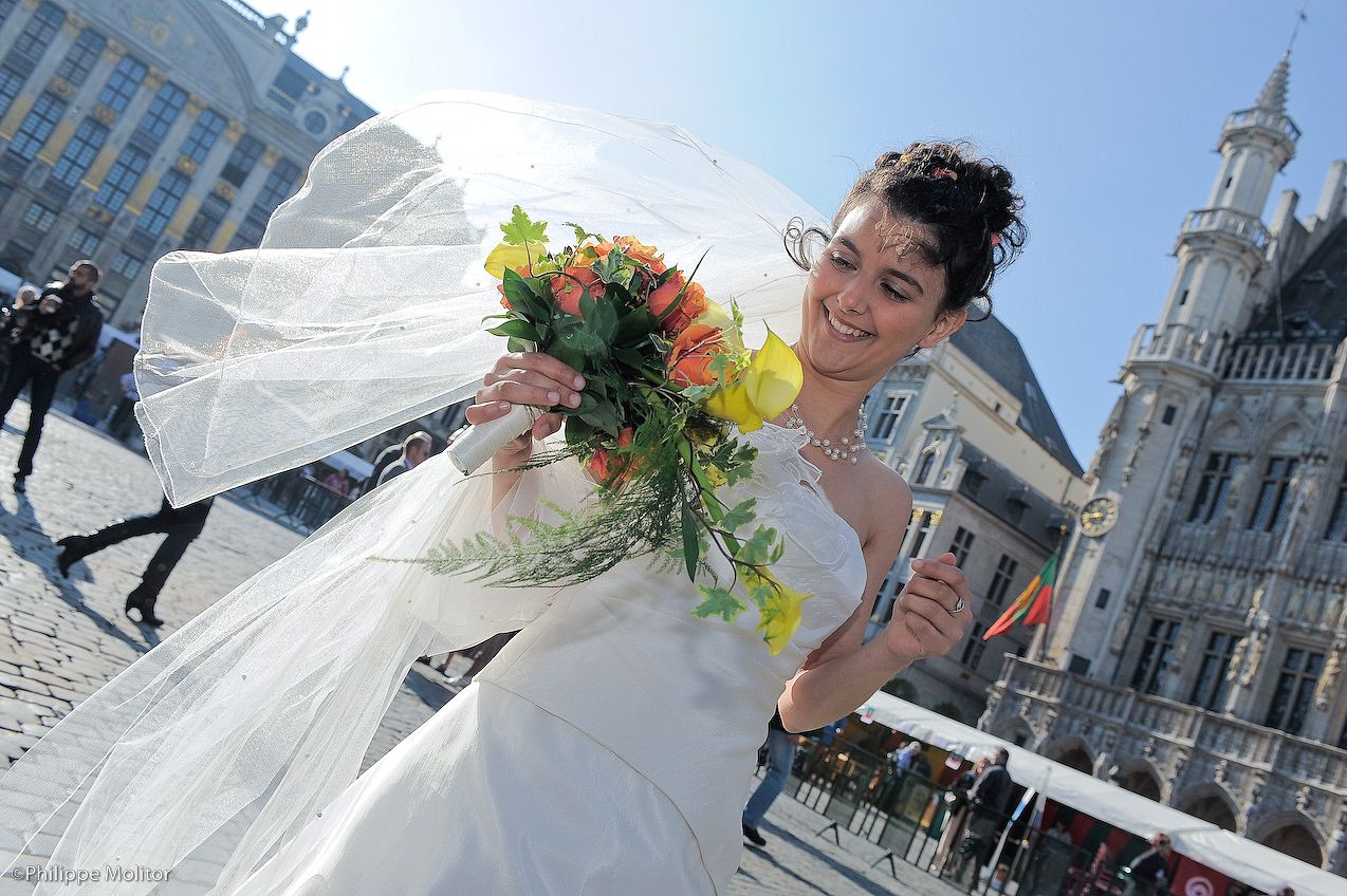 Smiling bride in white gown and veil holds colorful bouquet outdoors near historic European building on sunny day.