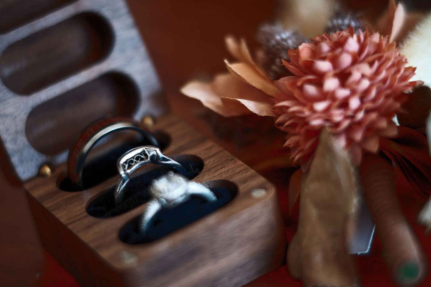 Two silver wedding rings displayed in an elegant wooden ring box beside a coral flower arrangement.