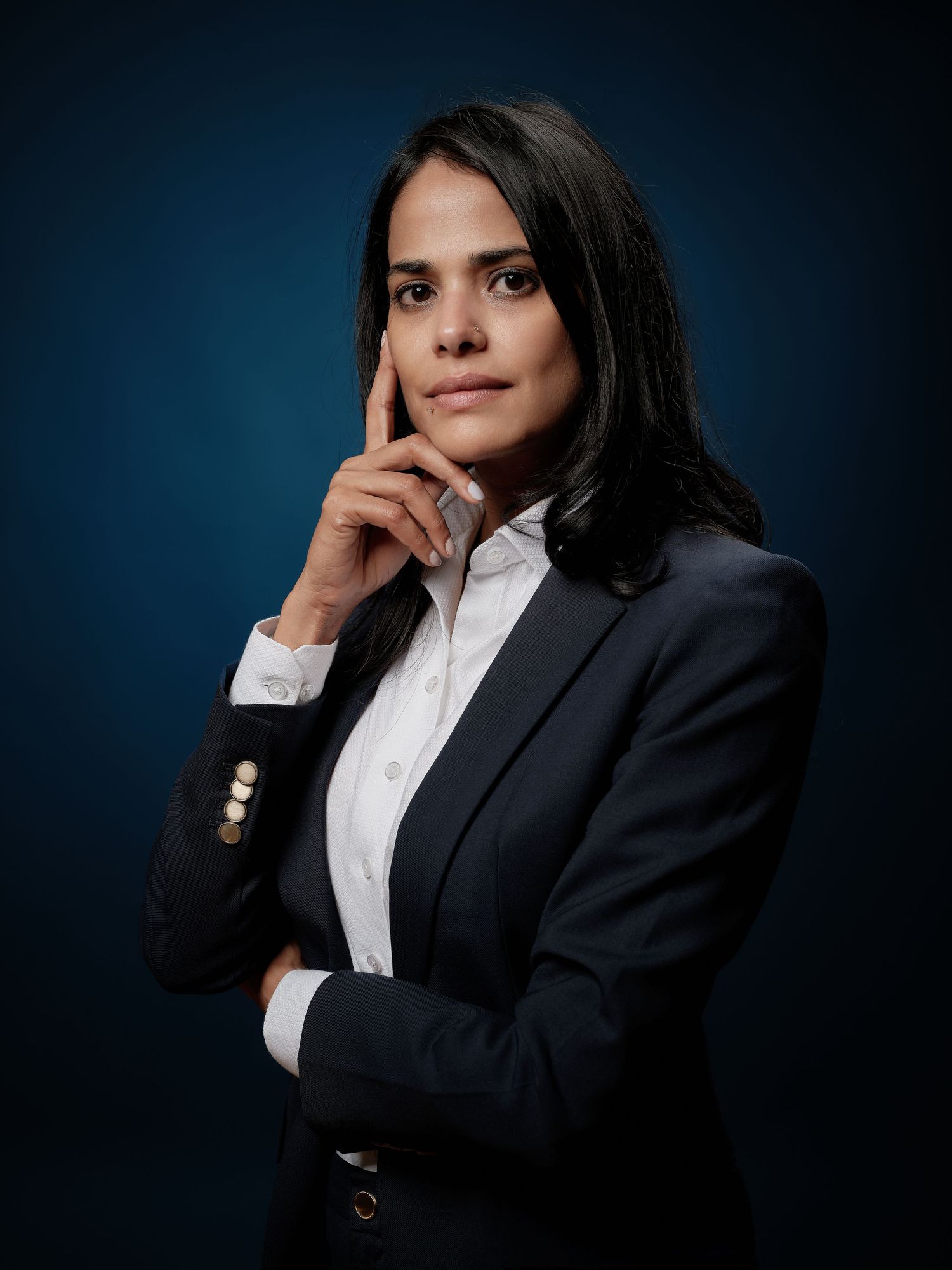 Professional woman in dark blazer poses thoughtfully against blue background in corporate portrait photo.