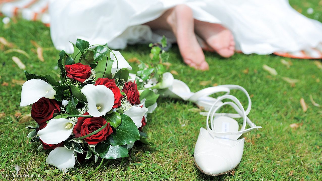 Bridal bouquet of red roses and white calla lilies on grass beside white wedding shoes with bare feet in background.