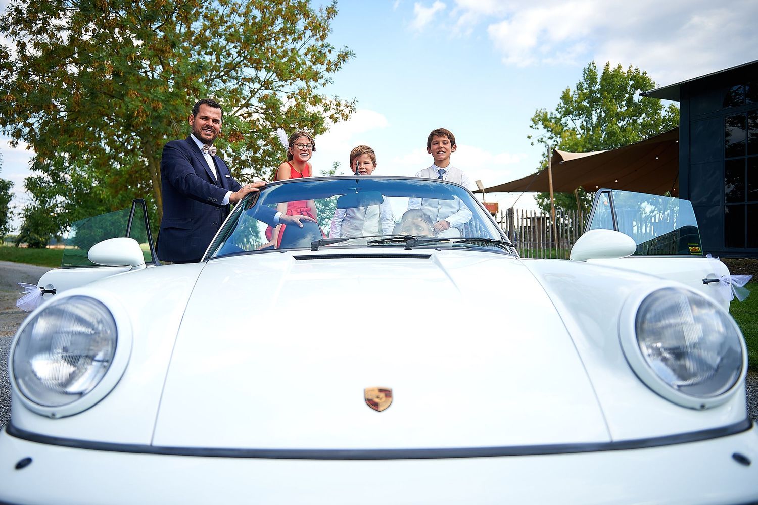 Four people posing with a classic white Porsche convertible outdoors on a sunny day.