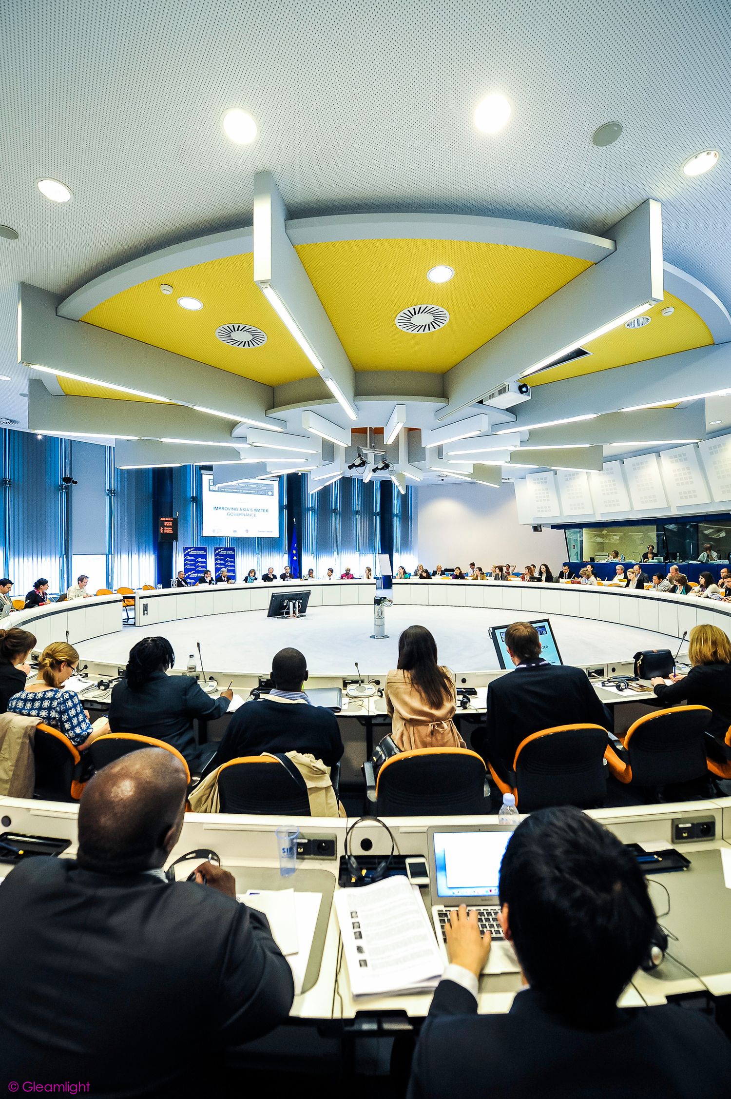Conference room with yellow ceiling panels and rows of attendees seated at tables.