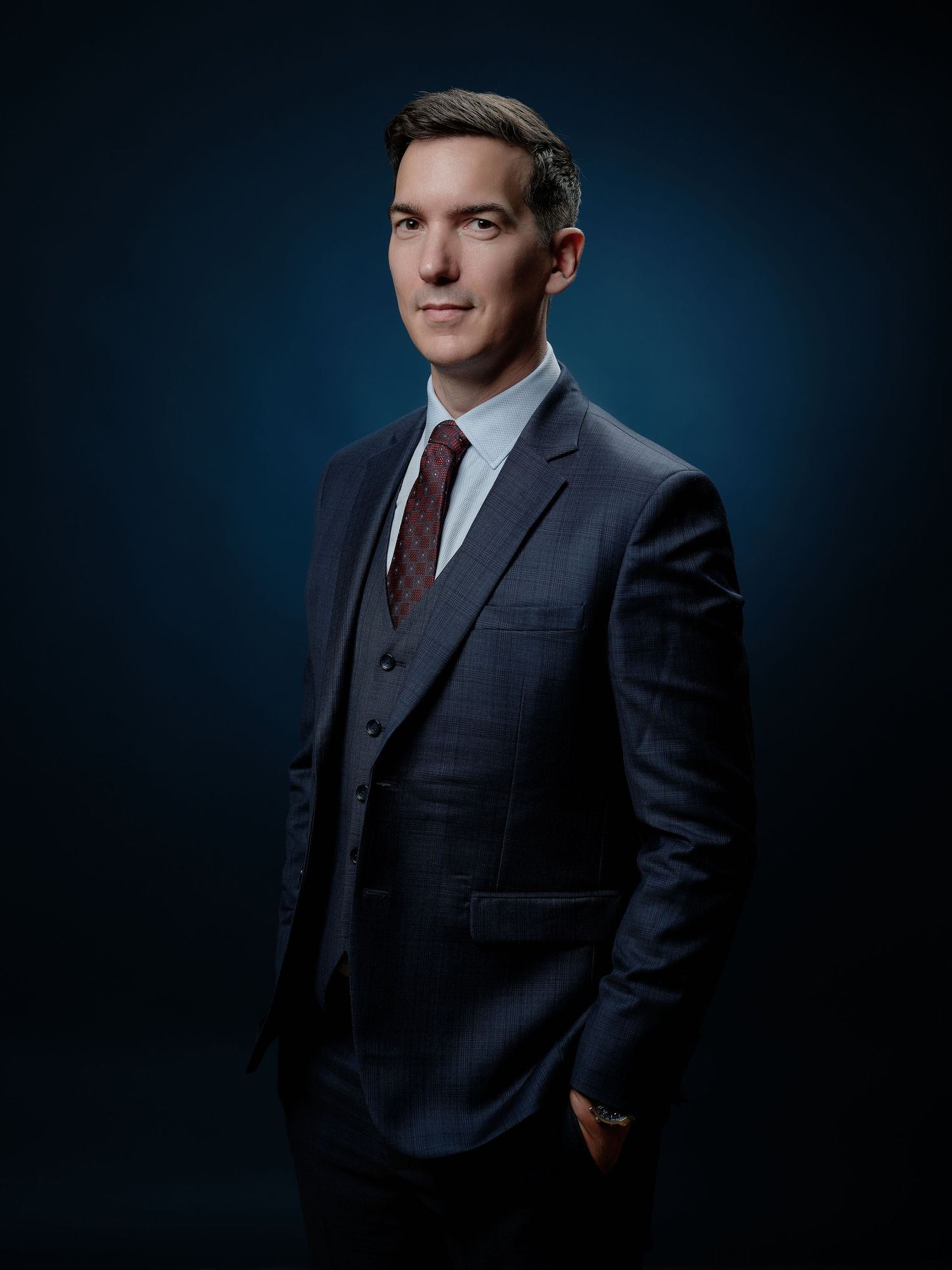 Professional man in navy blue three-piece suit and burgundy tie posing confidently against dark blue background. Portrait entreprise.