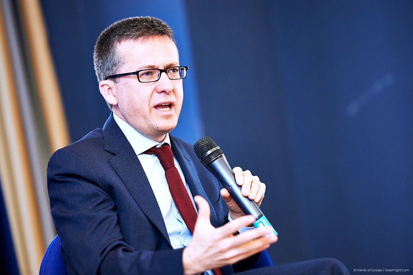 Speaker in business attire presenting at a corporate conference against blue backdrop.