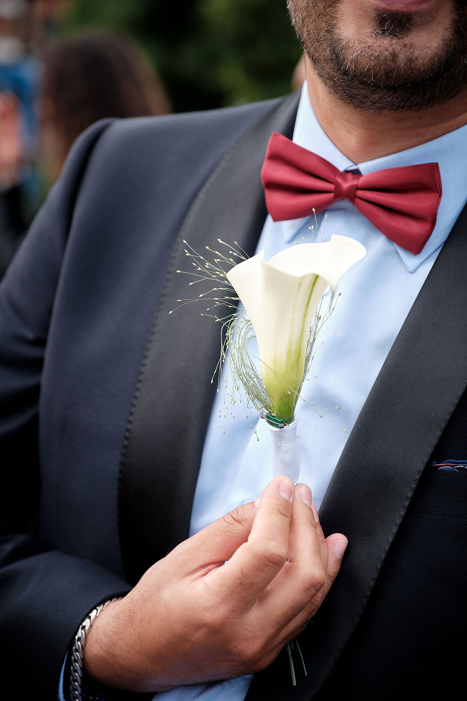 Groom in dark suit with red bow tie adjusting white calla lily boutonniere on wedding day.