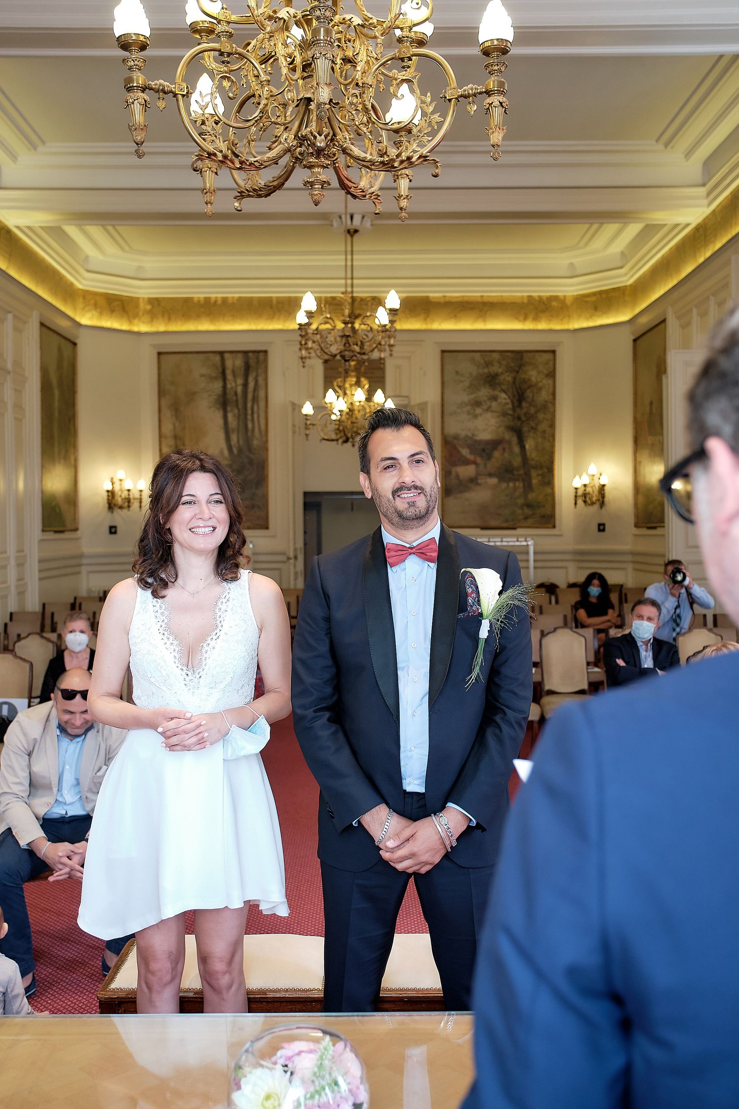 Couple stands at altar in elegant chandelier-lit hall during civil wedding ceremony, bride in white dress, groom in navy suit.