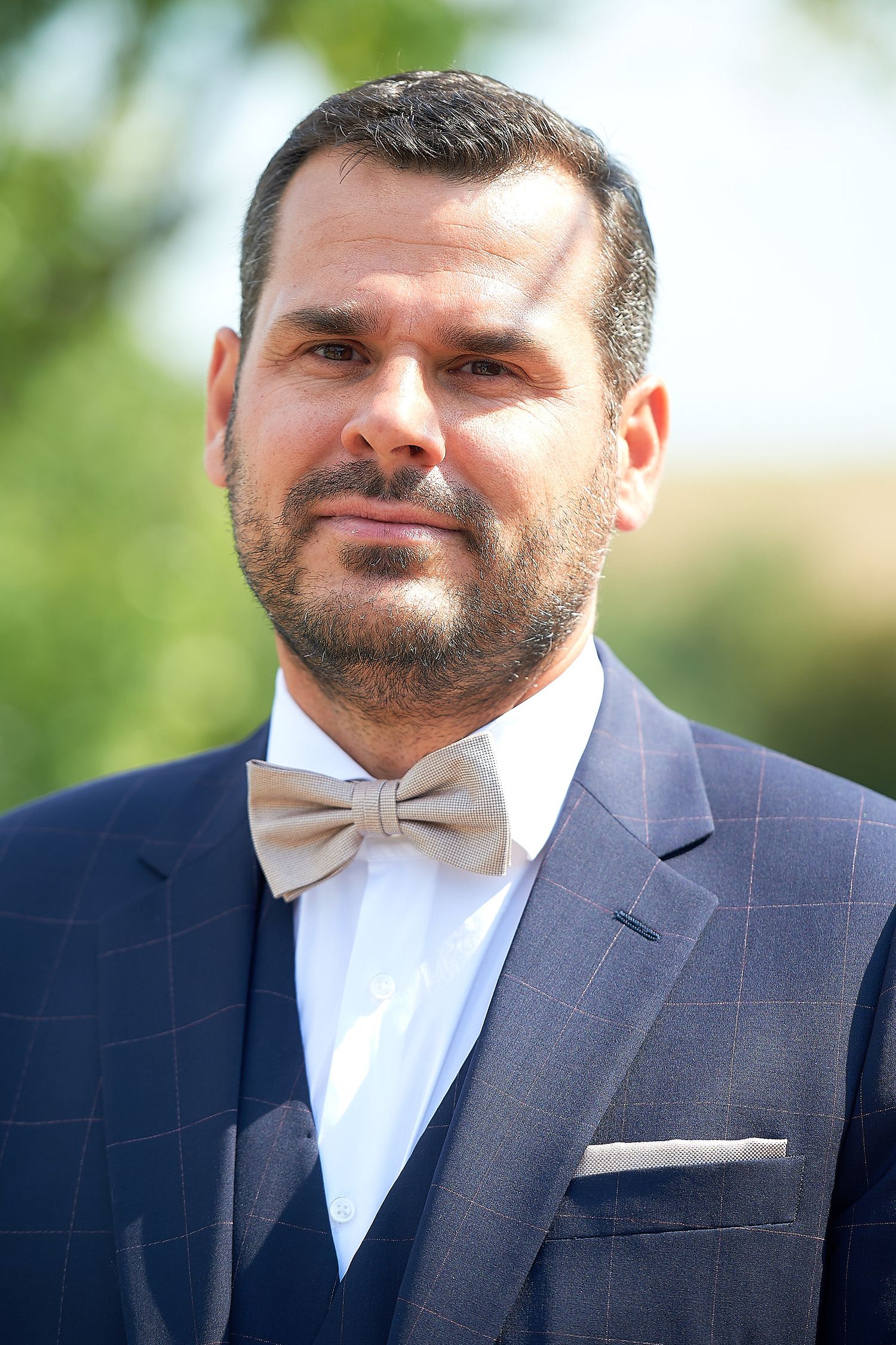 Man in navy plaid suit with beige bow tie and pocket square posing outdoors at formal event.