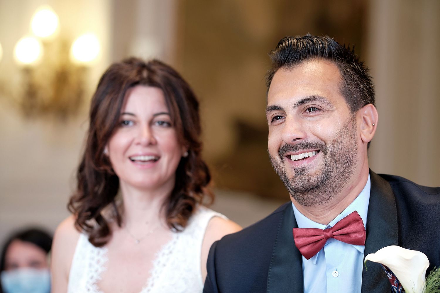 A smiling couple in wedding attire, man in bow tie and woman in white dress, at an indoor wedding ceremony.