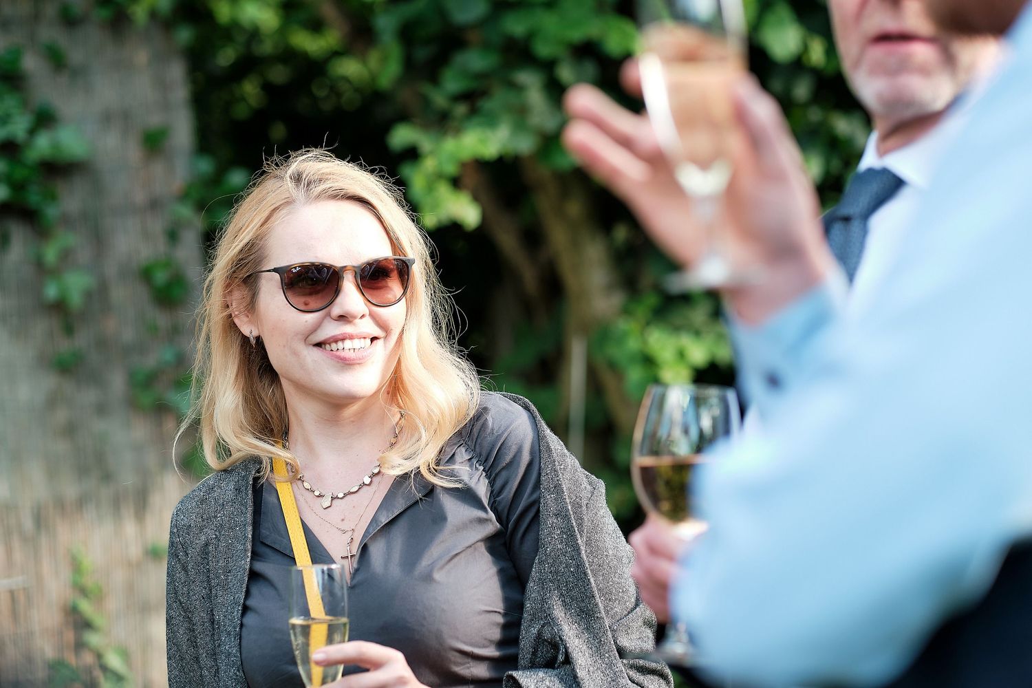 Smiling blonde woman in sunglasses enjoys a drink at an outdoor social gathering.