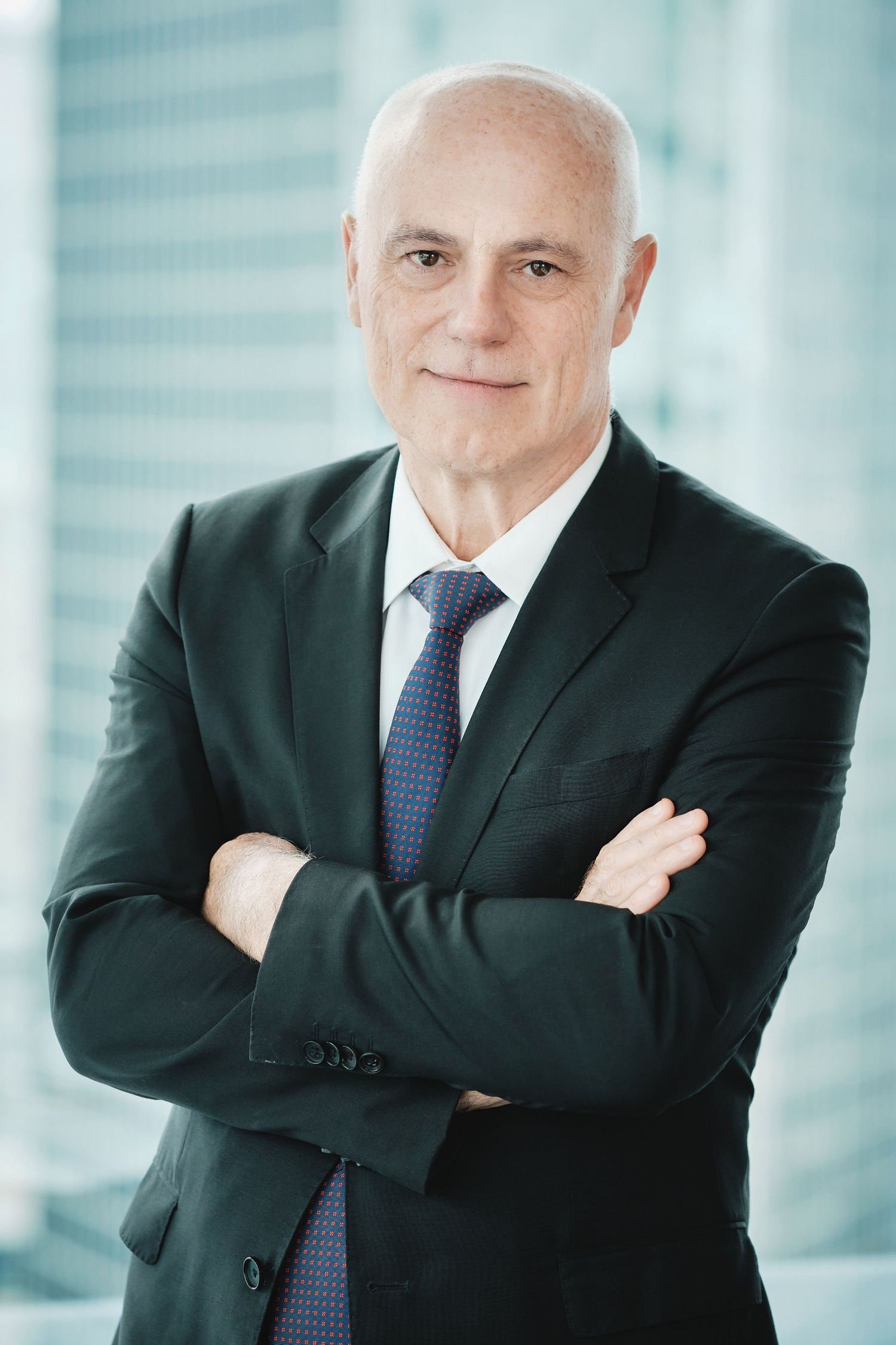 Corporate headshot of executive in dark suit with arms crossed against modern office setting.