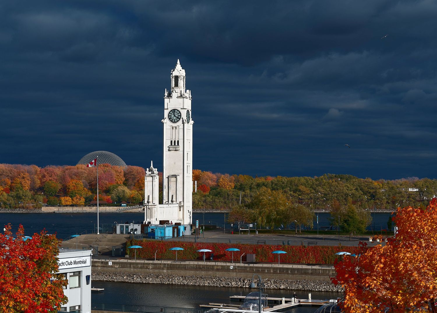 A panoramic view of a white church steeple against dark storm clouds with vibrant autumn foliage in the foreground.