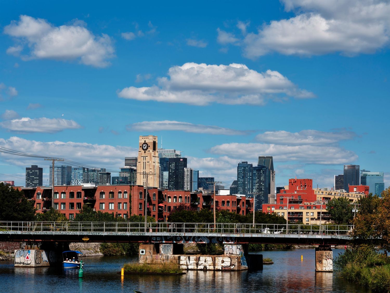 City skyline with brick buildings, clock tower, and modern skyscrapers reflected near a river bridge under a blue cloudy sky.