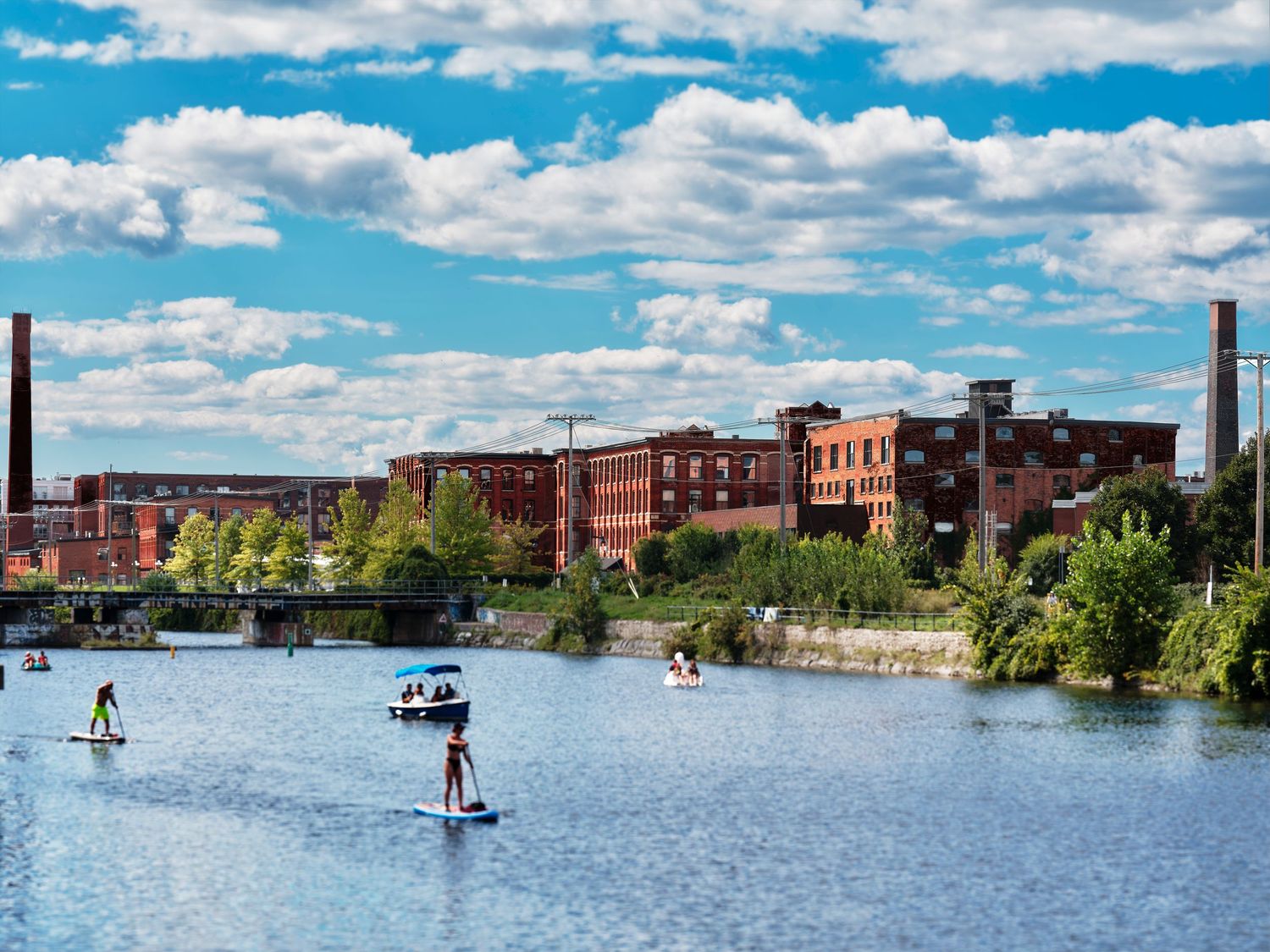 People paddle boarding on a river with historic red brick mill buildings and blue skies in the background.