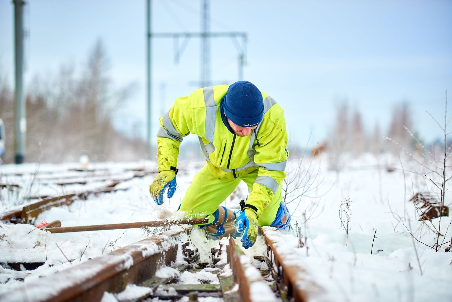 Railway worker in high-visibility clothing inspects track components in winter weather.