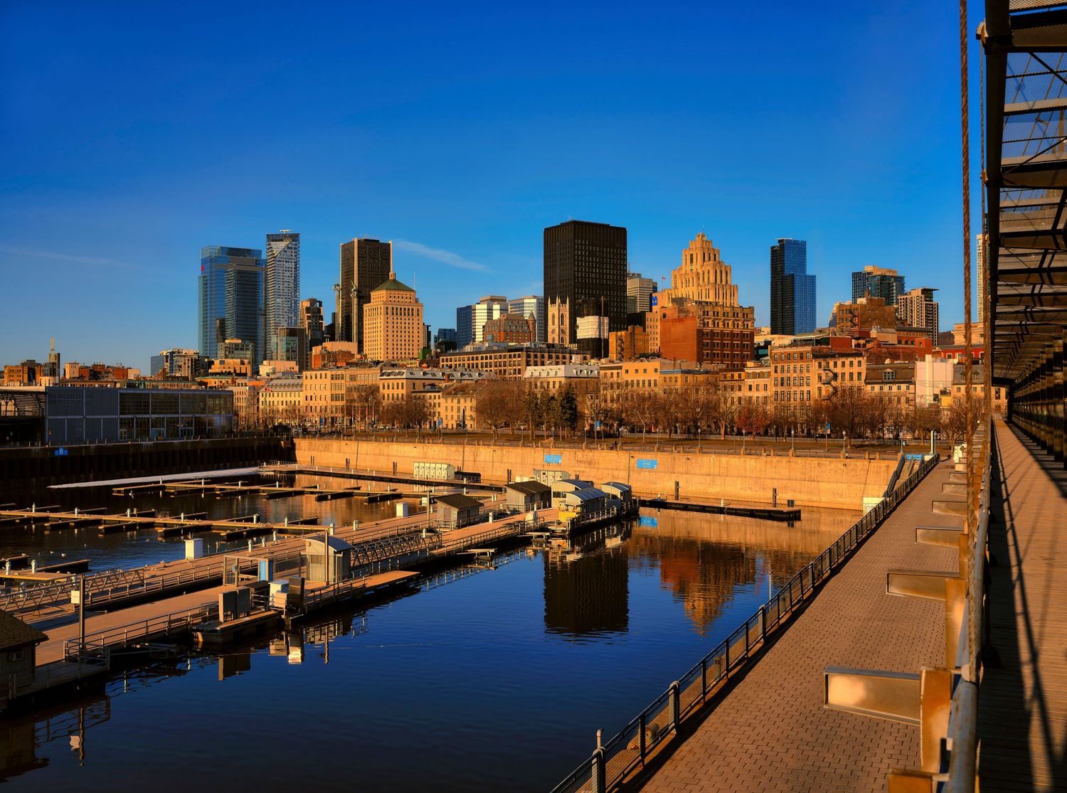 A panoramic view of Montreal's Old Port waterfront and skyline at sunset reflecting in the calm harbor waters.