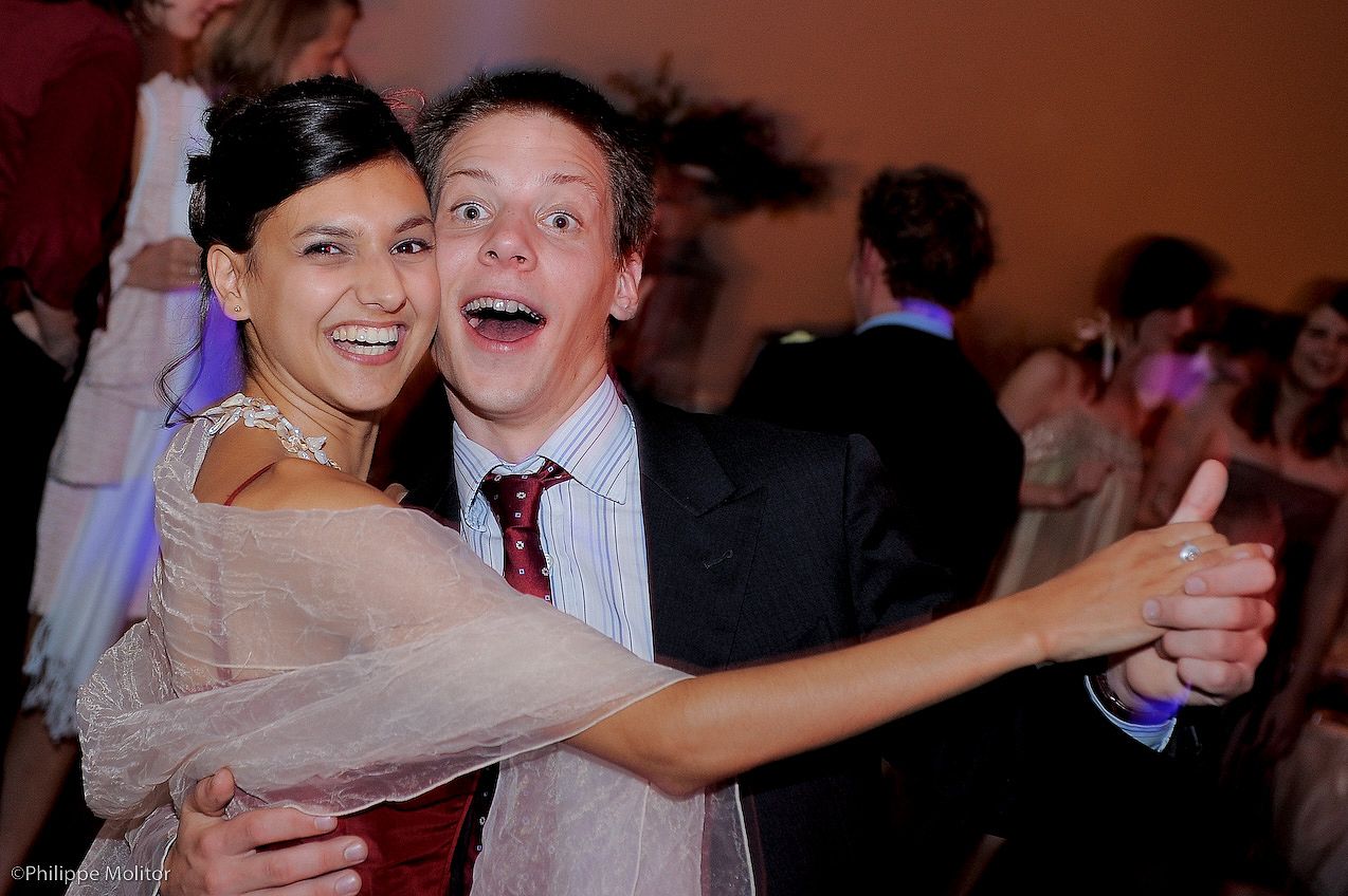 A smiling couple dancing at a formal event, woman in silver dress and man in suit giving thumbs up.