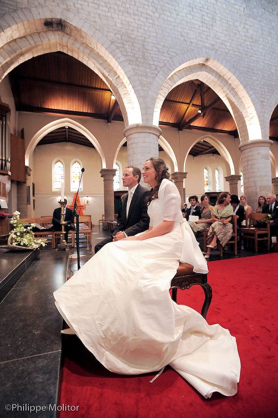 Bride in white gown and groom seated at church altar during wedding ceremony with red carpet aisle and stone arches.