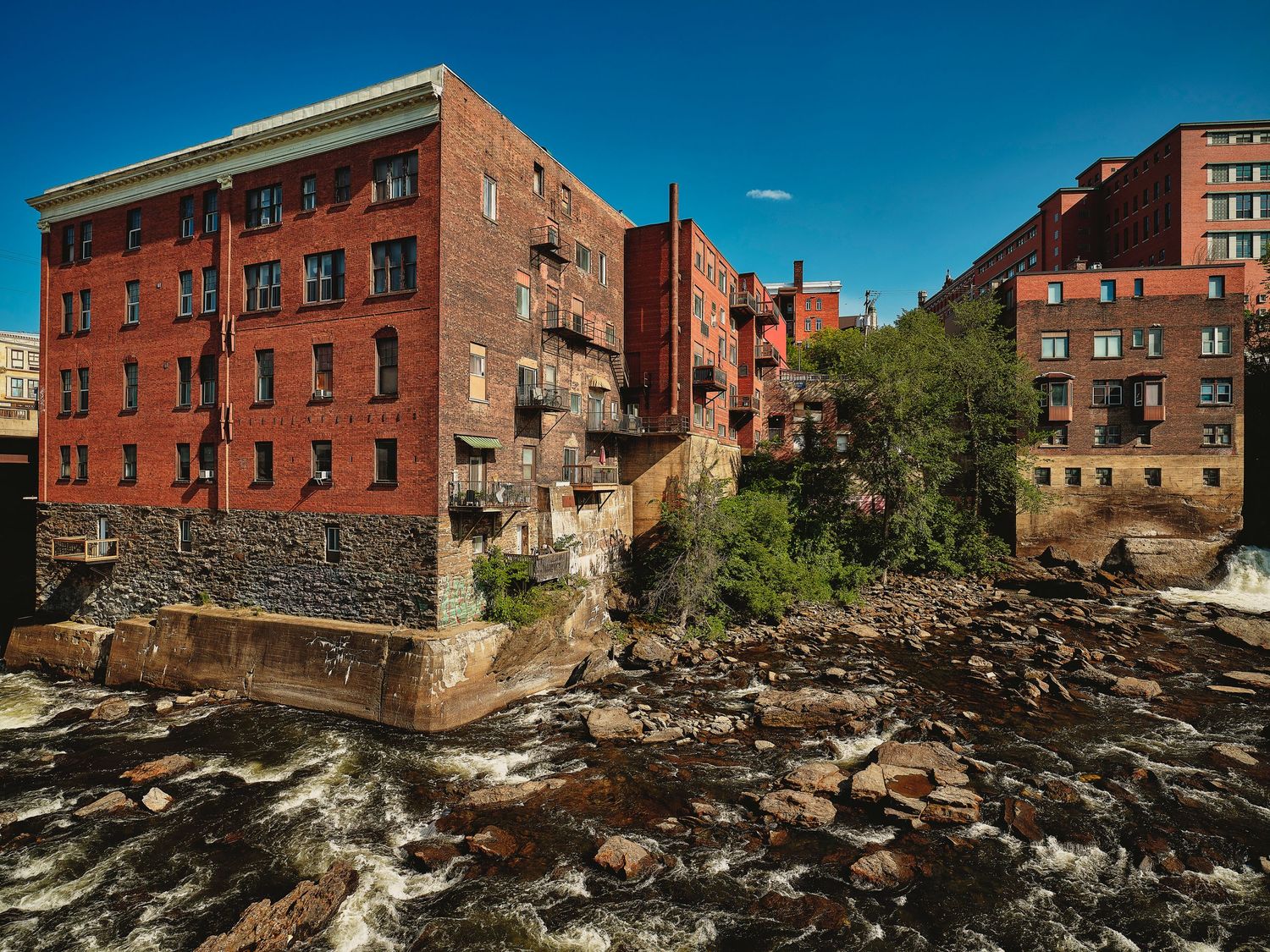 Historic red brick industrial buildings along rocky riverbank under clear blue sky.