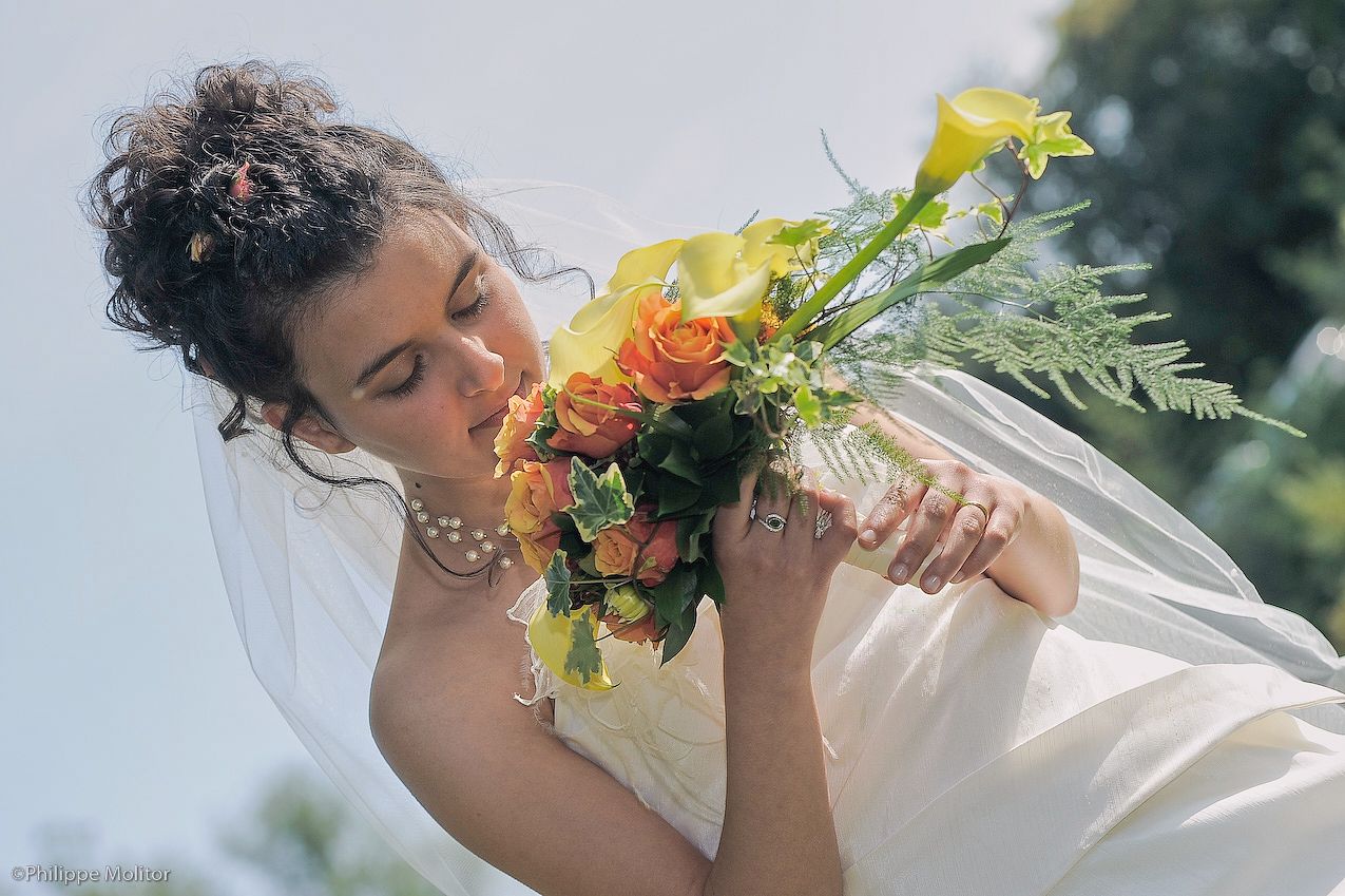 Bride in white gown and veil smelling a vibrant orange and yellow wedding bouquet outdoors on her wedding day.
