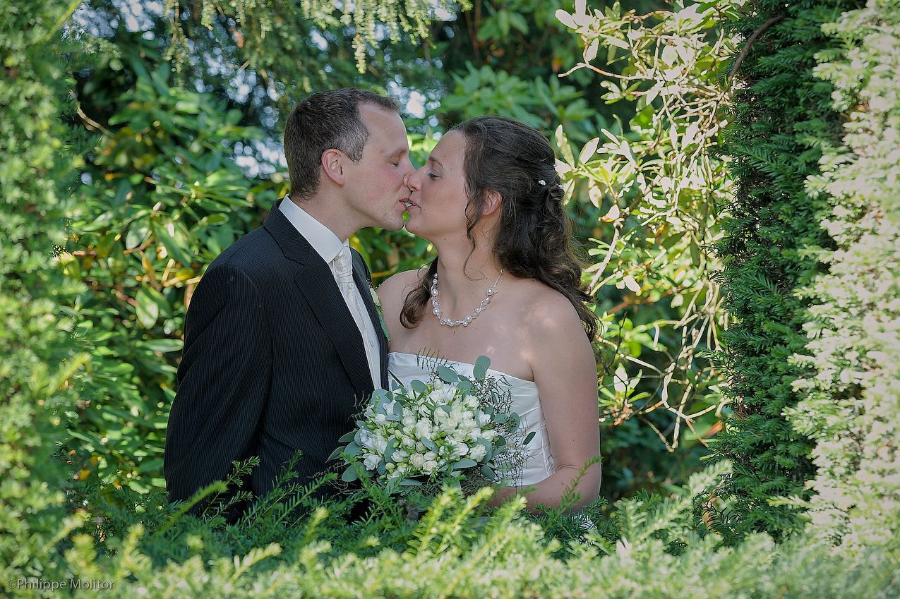 A bride and groom share a romantic kiss surrounded by lush green foliage, bride holding a white bouquet.