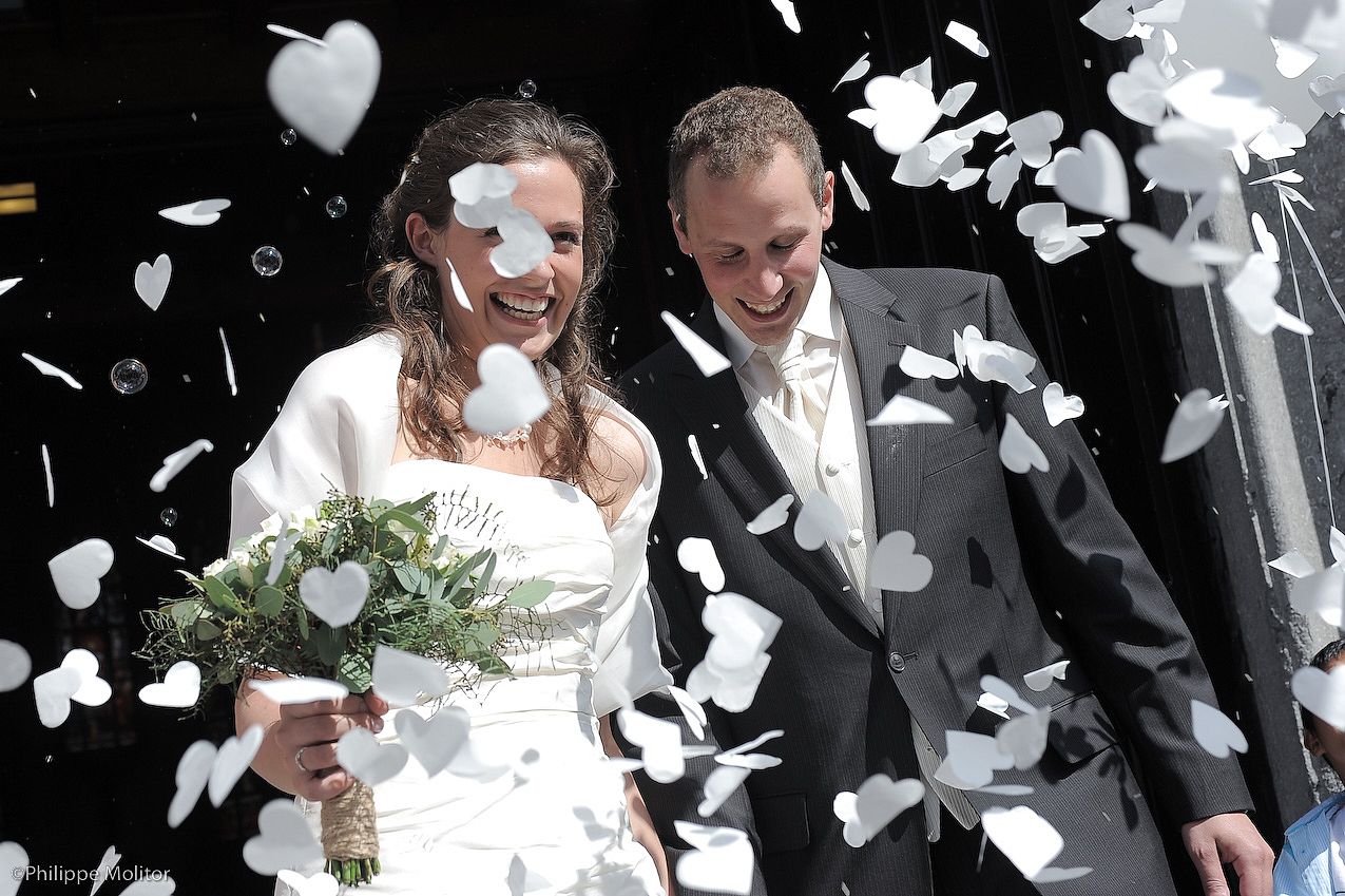 Joyful newlyweds celebrate outside church surrounded by white heart-shaped confetti, bride holding bouquet.