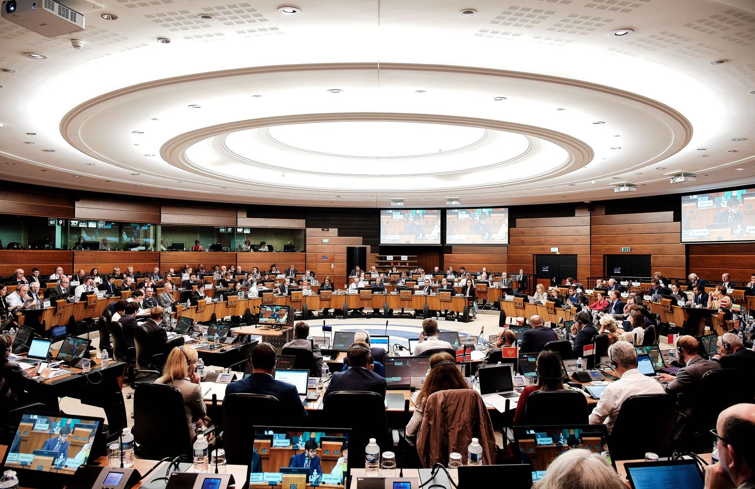 Wide panoramic view of a large conference hall with circular lighting and rows of attendees seated at tables.