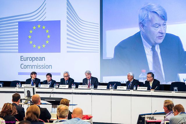 European Commission meeting room with EU flags and official branding displayed.