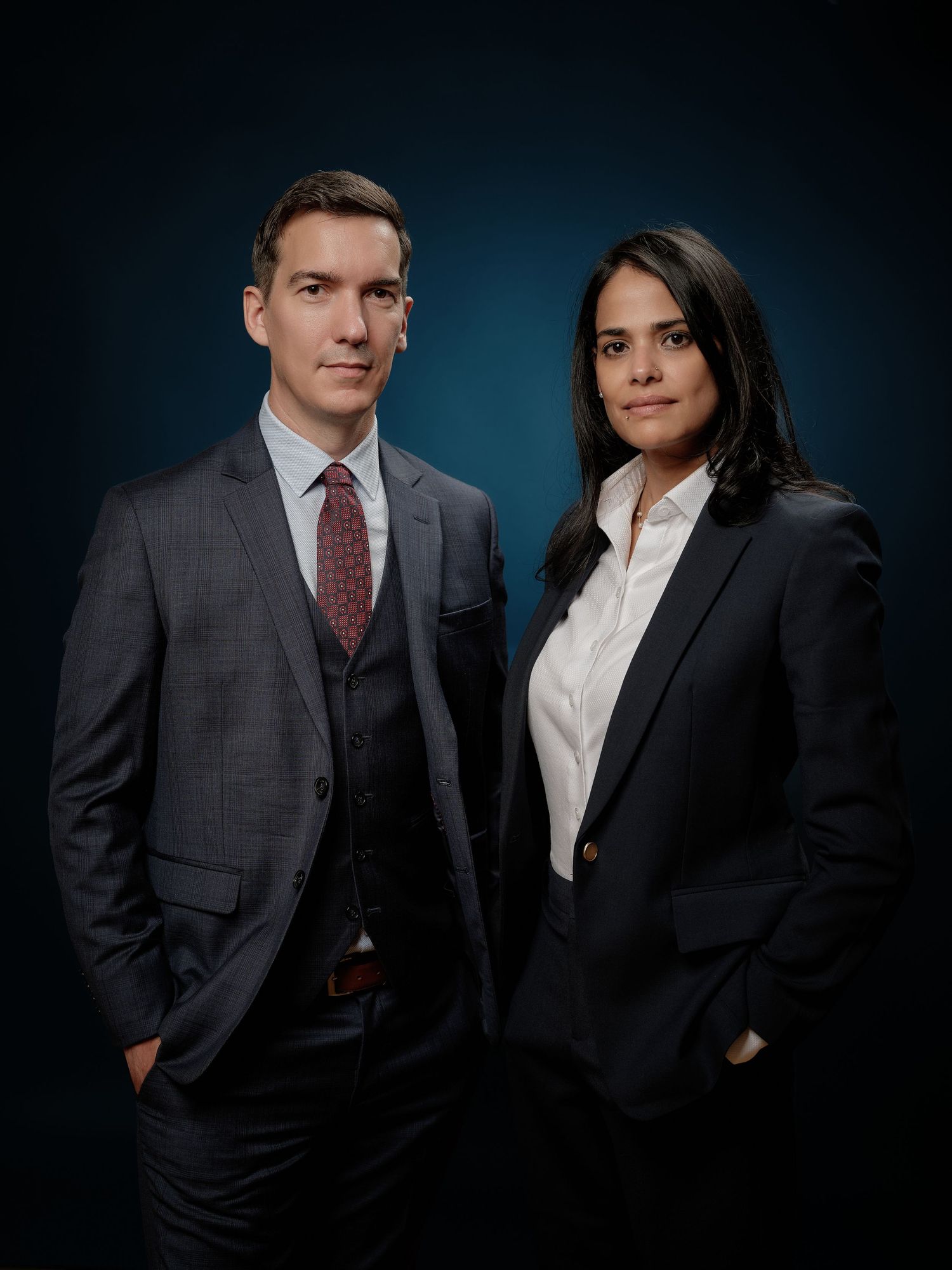 Two professionals in formal business attire pose for a studio portrait against a dark blue background.