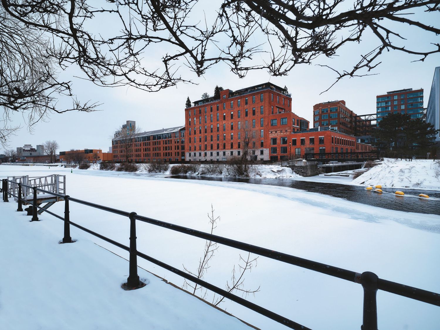 Snow-covered riverbank with industrial buildings and warehouses along the winter waterfront.