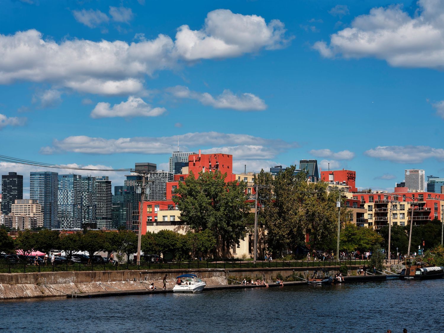 Waterfront cityscape with colorful buildings, lush trees, and a blue sky with white clouds reflecting on calm water.