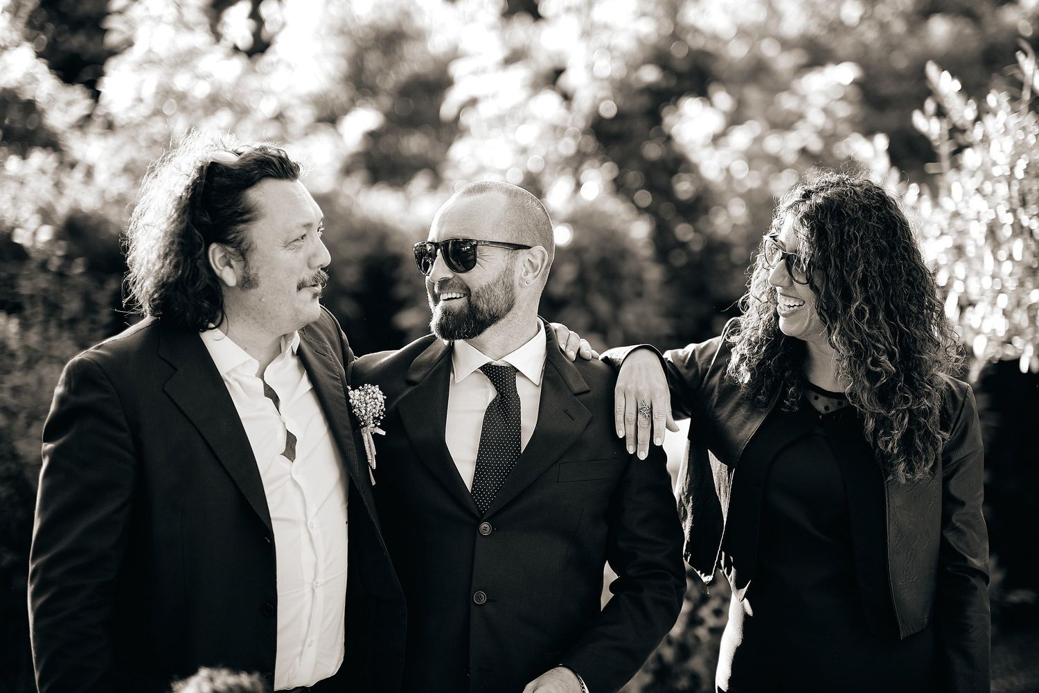 Three men in black suits smiling together outdoors in black and white wedding photo.