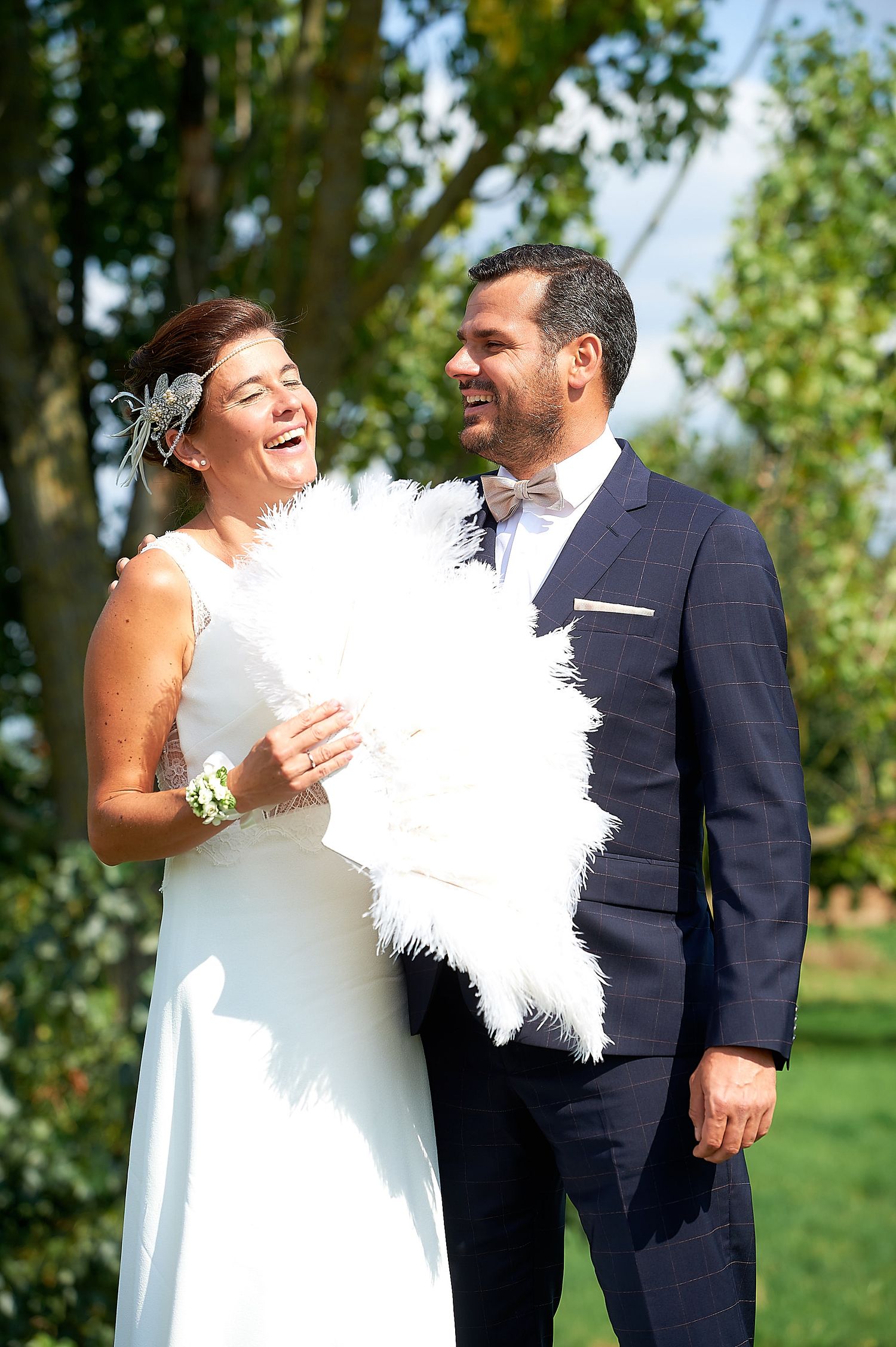 Happy bride and groom laughing outdoors, bride holding white feather fan in elegant wedding attire.
