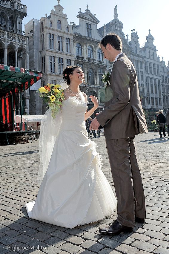 Bride in white gown holds yellow bouquet while groom in gray suit holds her hand at Grand Place, Brussels.