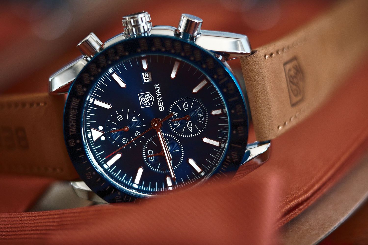 TAG Heuer chronograph watch with blue dial and brown leather strap resting on a tan surface.