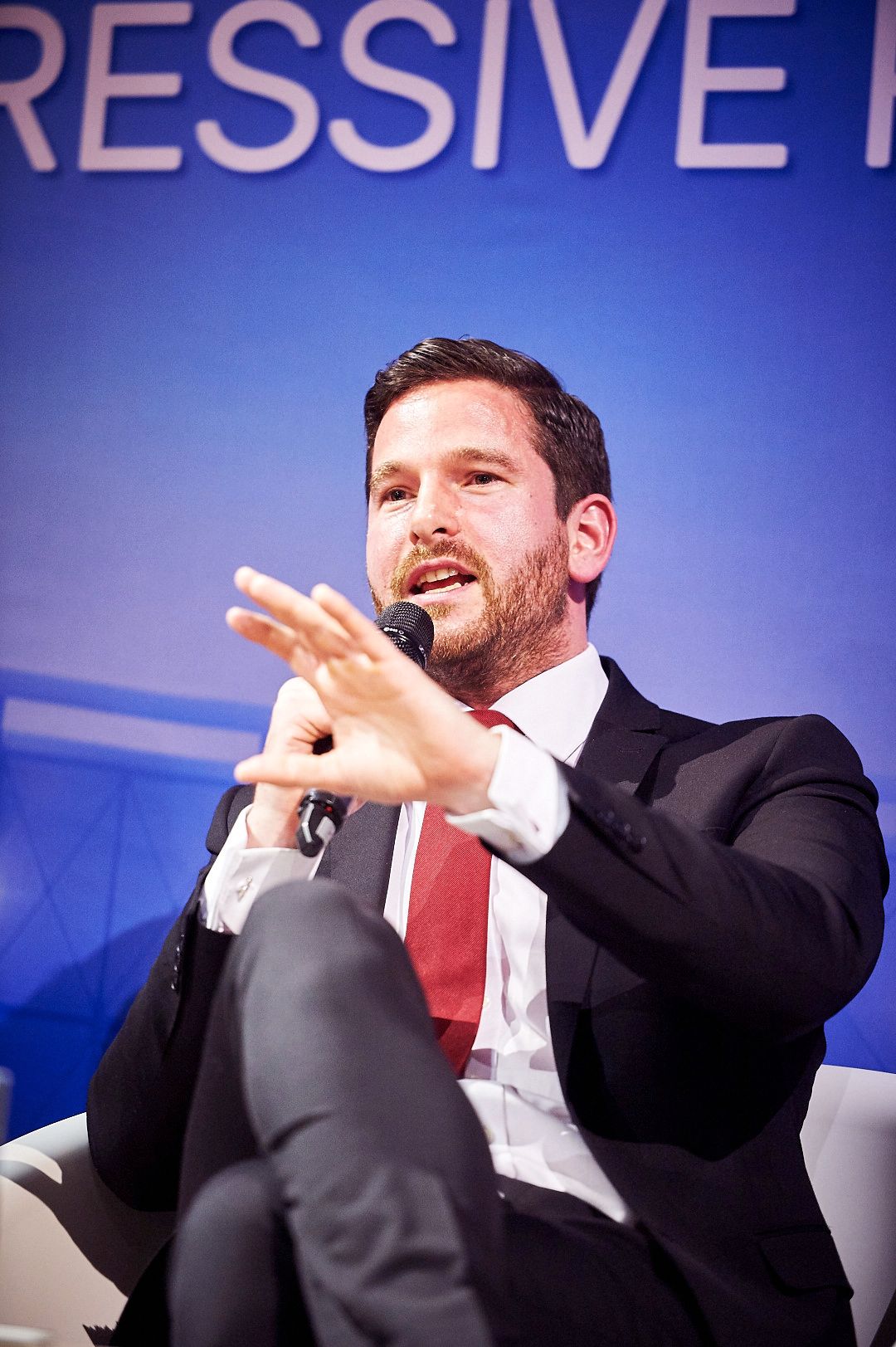 Professional speaker in business attire gesturing during presentation against blue background.