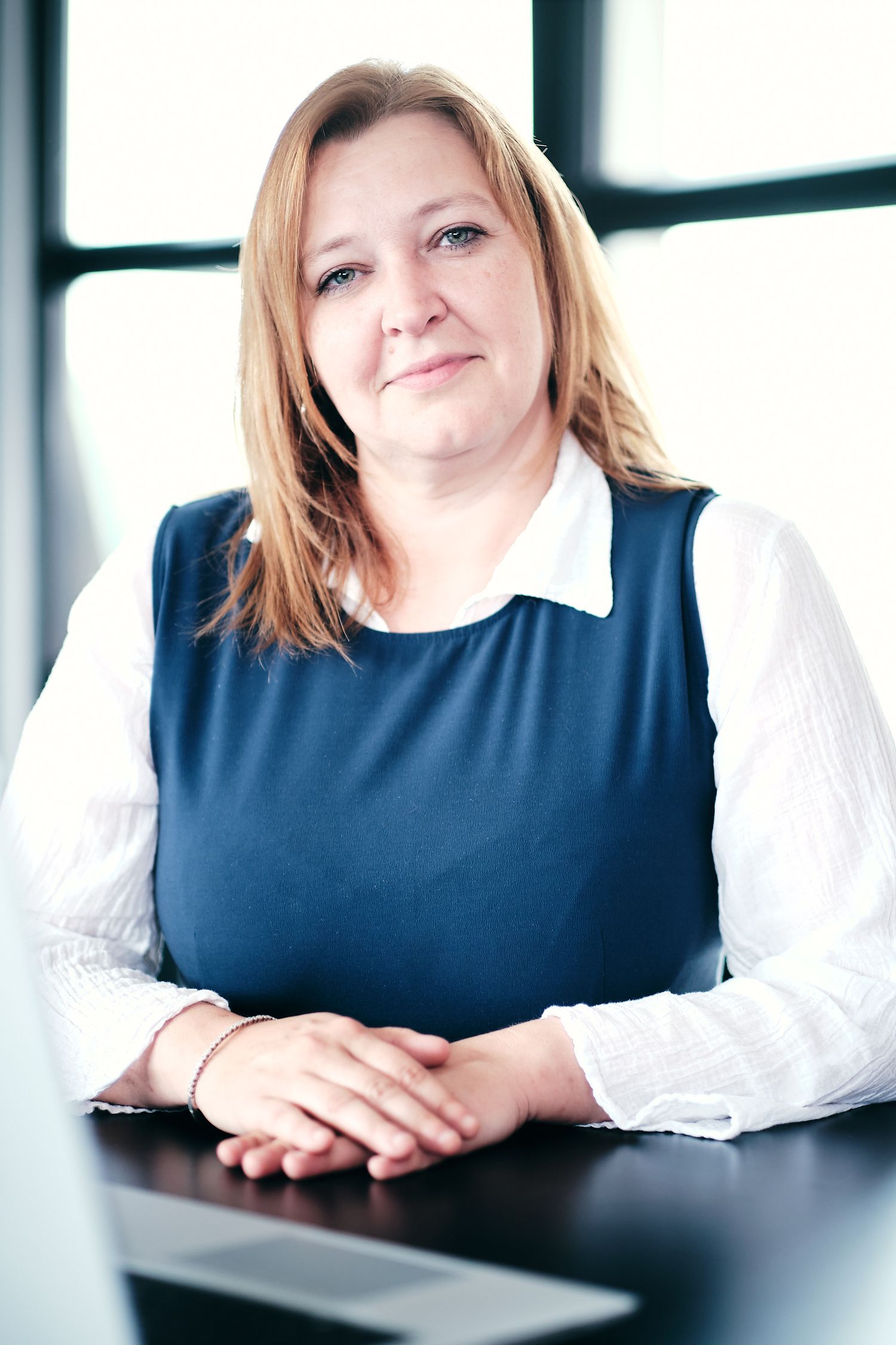 Business professional in navy sleeveless dress and white blouse seated at desk in modern office.