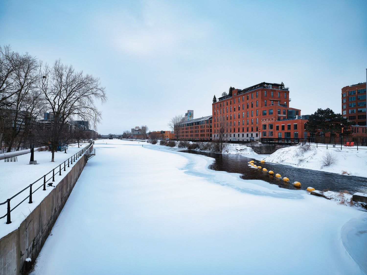 Snow-covered riverbank with historic brick buildings and bare trees in winter panorama.