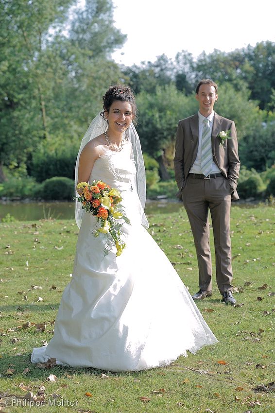 Bride in white gown holding colorful bouquet stands before groom in brown suit outdoors.