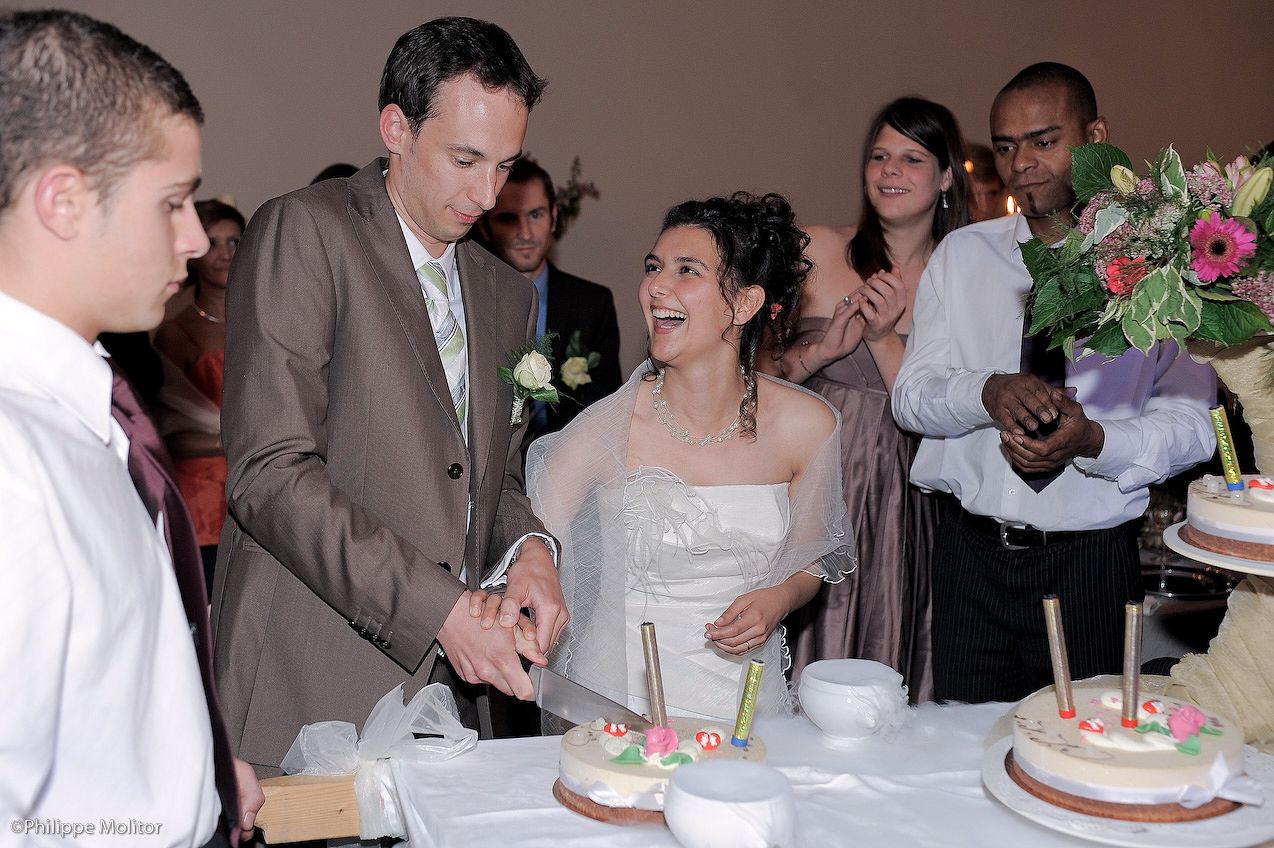 Bride and groom cutting wedding cake together surrounded by smiling guests at elegant reception.