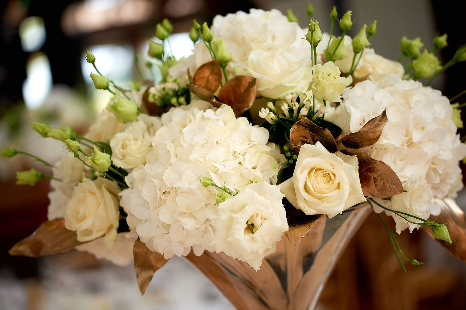 Elegant white hydrangea and rose floral centerpiece with brown leaves in a vase for a wedding reception.