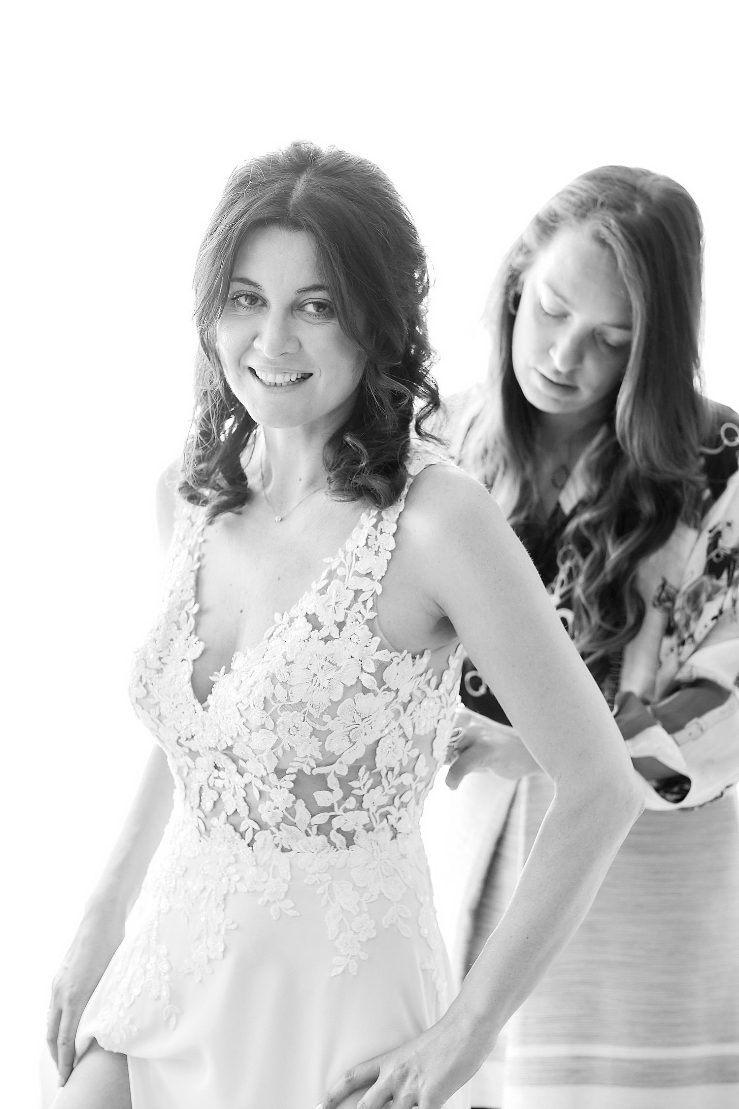 Black and white photo of a bride in lace wedding dress smiling as bridesmaid helps fasten her gown.
