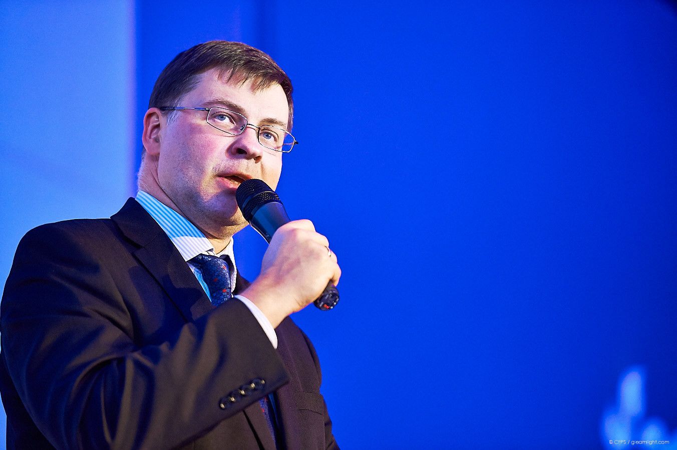 Speaker in a dark suit presenting against a blue background at a conference.