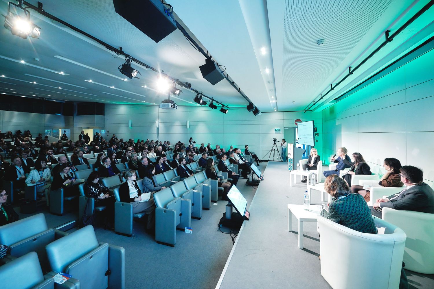 Modern conference hall filled with attendees under blue and green lighting during presentation.