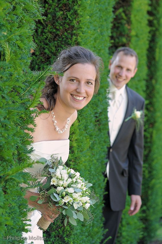 Smiling bride holding white bouquet with groom in gray suit standing among lush green hedges at wedding.