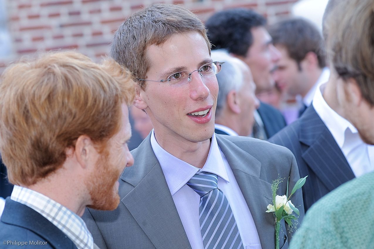 A young man in a gray suit with a boutonniere smiles during a wedding reception outdoors.
