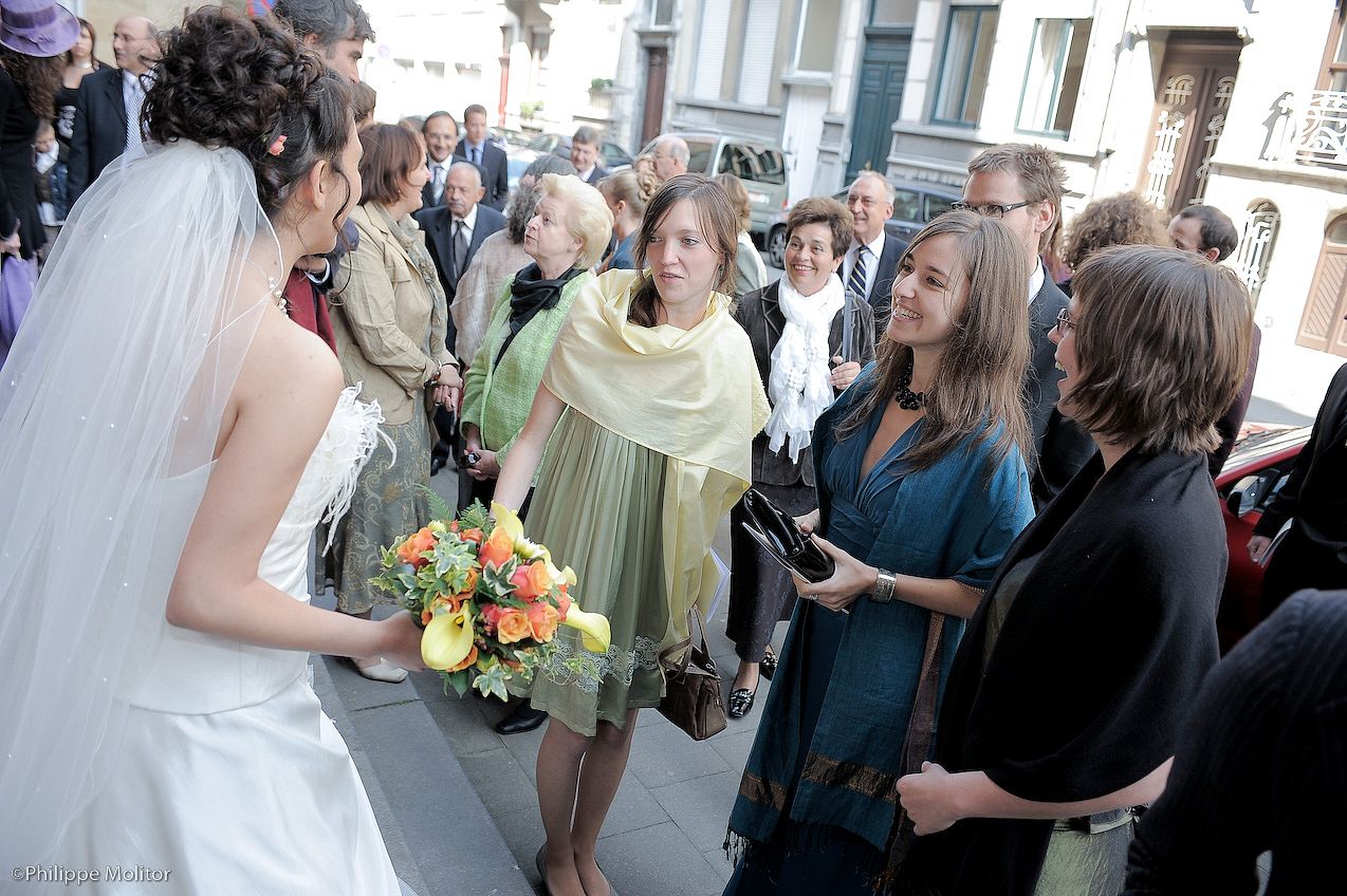 Bride in white gown with veil greeting guests outside church, holding vibrant orange and yellow bouquet.
